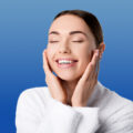 Woman in white sweater holding her face while smiling with eyes closed on blue background